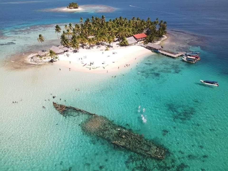 Aerial view of San Blas Islands beach with clear turquoise waters, palm trees, and people snorkeling near a rocky outcrop, captured by Bandits Adventure Tours Panama.