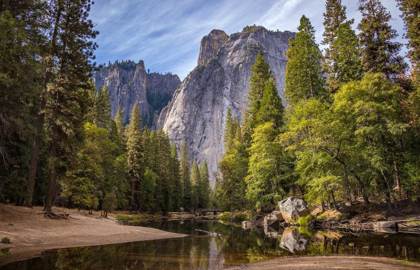 Towering granite cliffs rise above a calm river surrounded by lush evergreen trees in a serene natural setting.