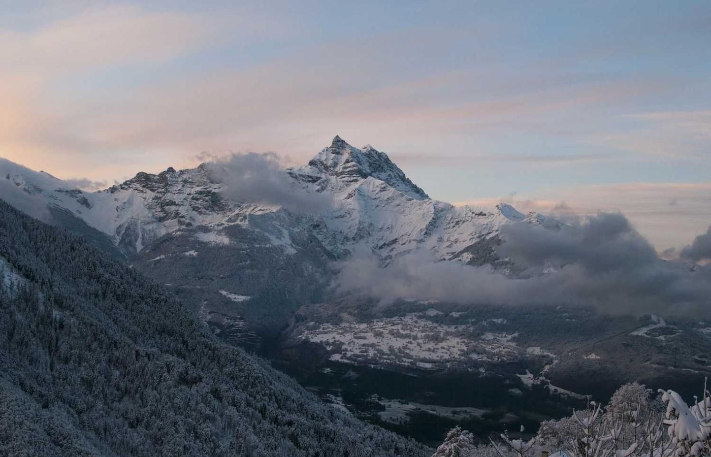 Snow-capped mountain peak surrounded by clouds, with a forested slope and valley settlement below during dawn or dusk.