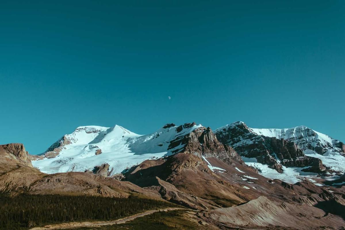Snow-capped mountains rise majestically against a clear blue sky, with a dirt path winding through a forested area below.