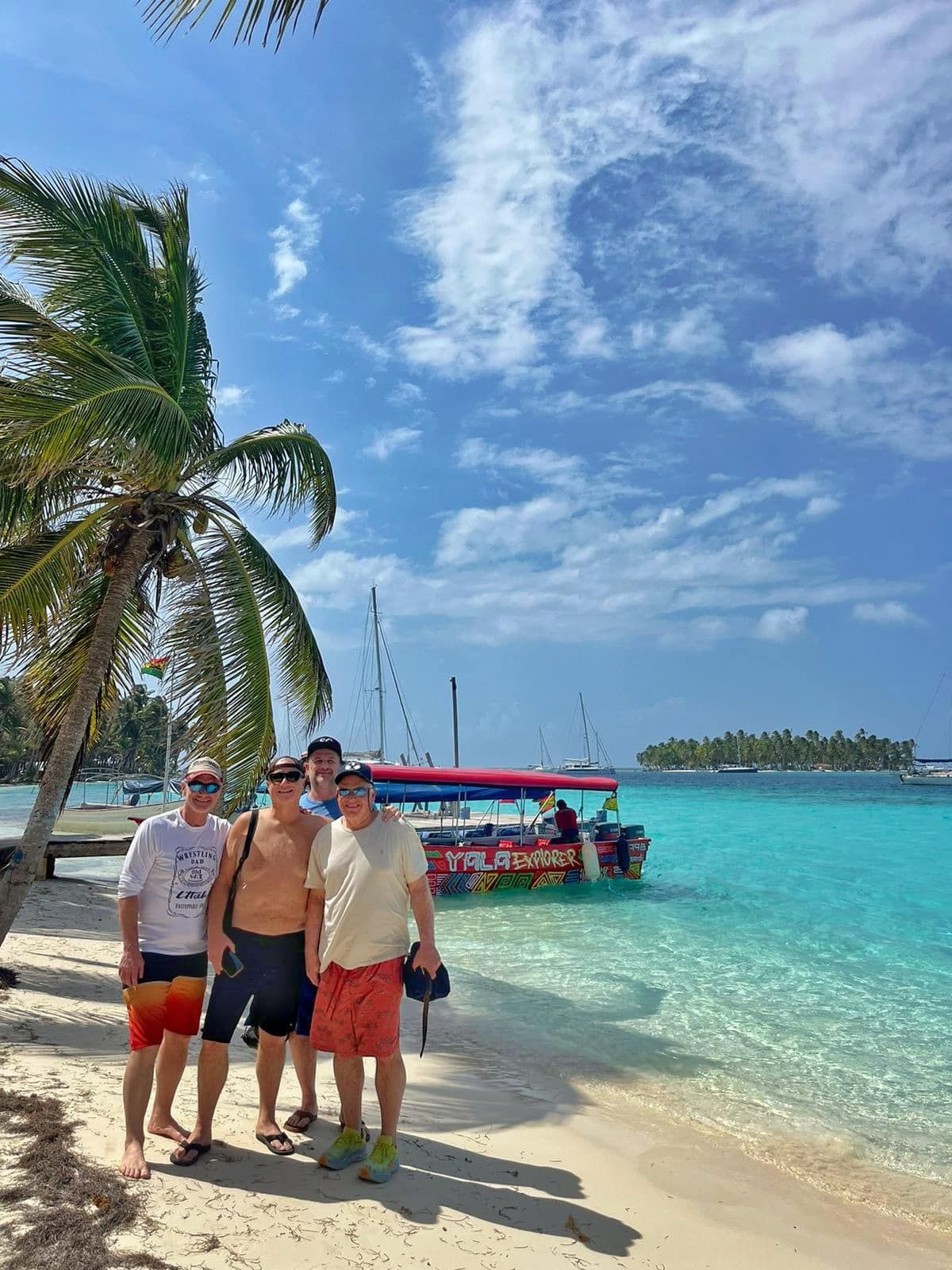 Four men enjoying a sunny day on a tropical beach during the San Blas Islands Day Tour Guna Yala in Panama City, with a colorful boat and turquoise waters in the background.