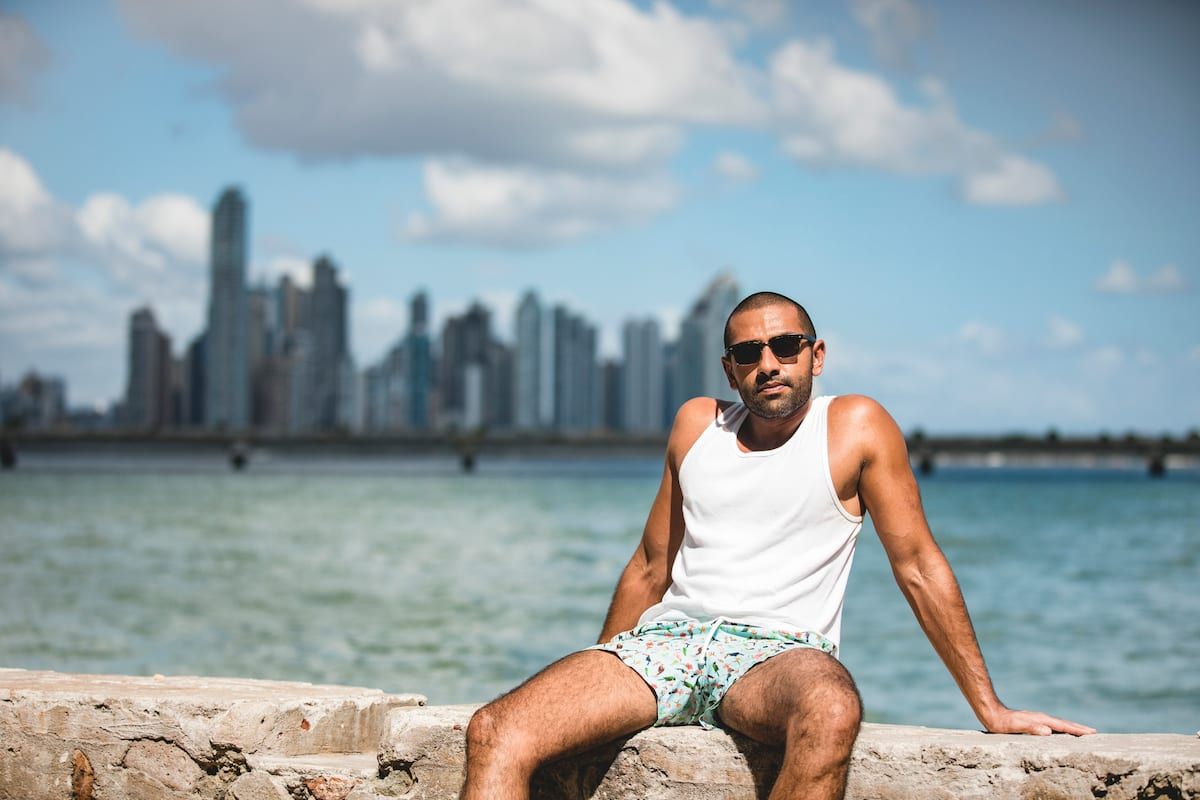 Man in white tank top and shorts sits on stone wall overlooking ocean with Panama City skyline in background during Casco Antiguo Photo Walking Tour