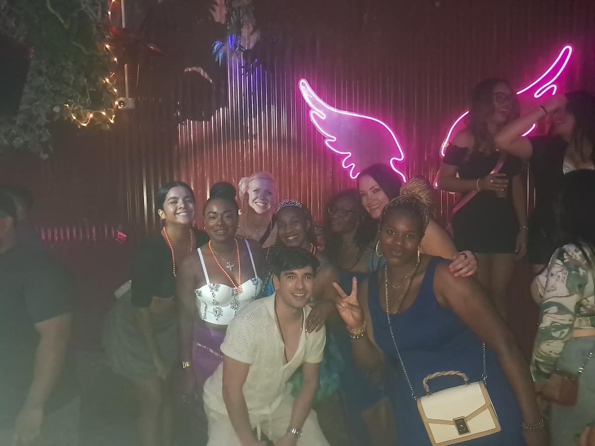 Group of friends posing for a photo in front of a neon angel wing sign during a bar crawl in Casco Viejo, Panama.