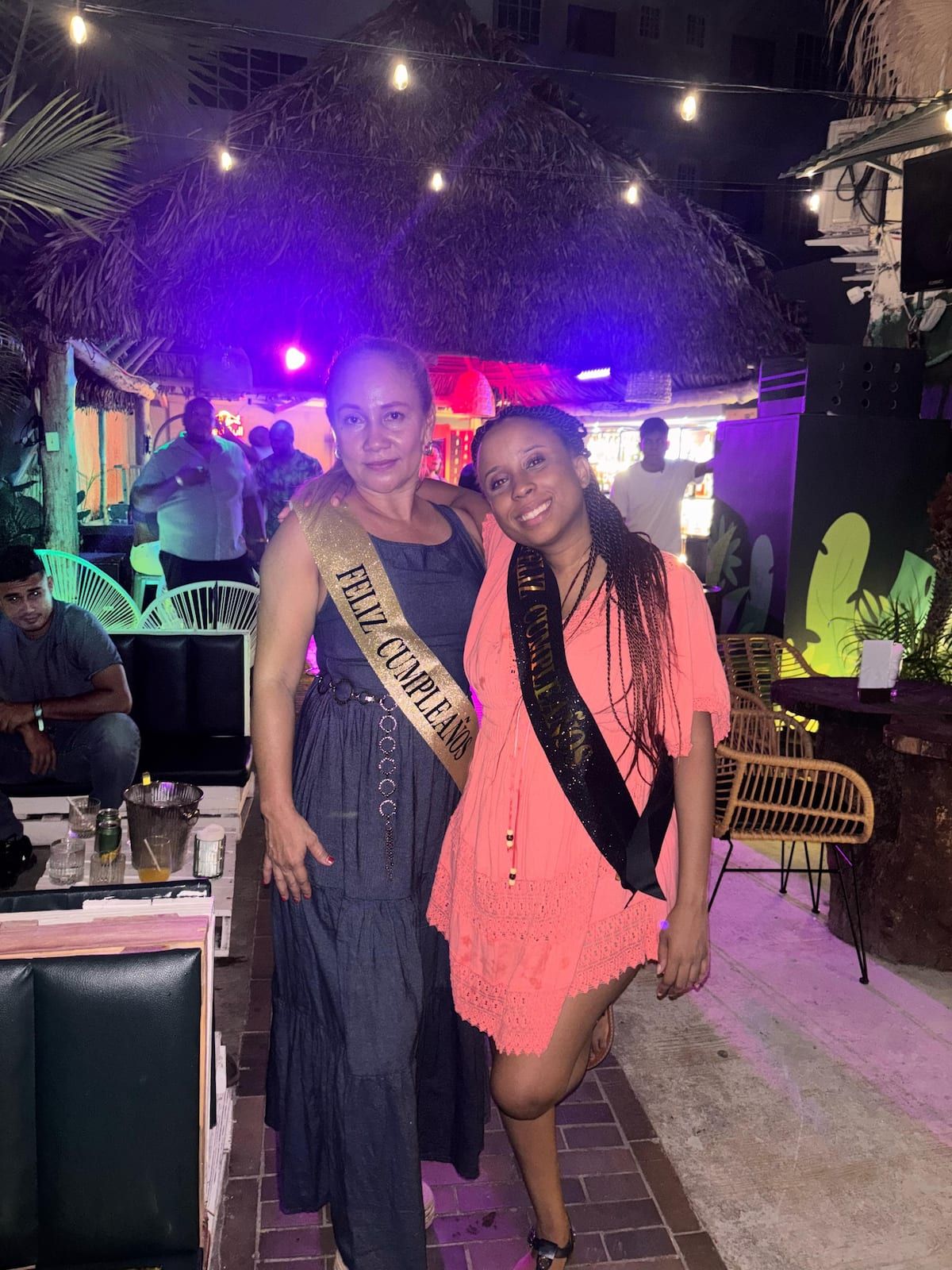 Two women celebrating a birthday at a lively bar crawl event in Casco Viejo, Panama City, wearing 'Feliz Cumpleaños' sashes under colorful string lights.