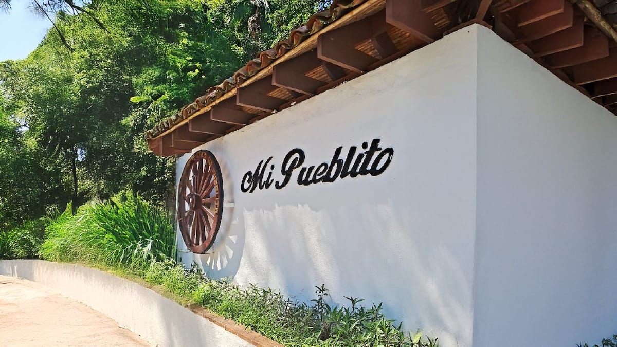Pared blanca con el texto 'Mi Pueblito' y rueda de carro de madera en un jardín tropical durante una caminata en Cerro Ancon en la Ciudad de Panamá