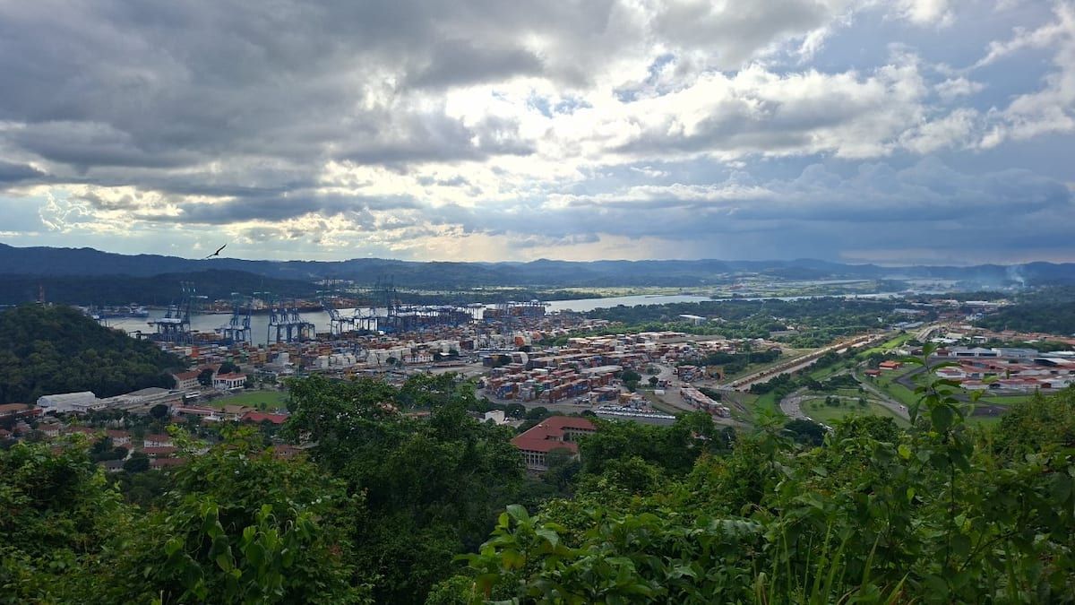 Vista panorámica desde la caminata del Cerro Ancón en la Ciudad de Panamá, con el puerto, grúas de contenedores y exuberante vegetación bajo un dramático cielo nublado.