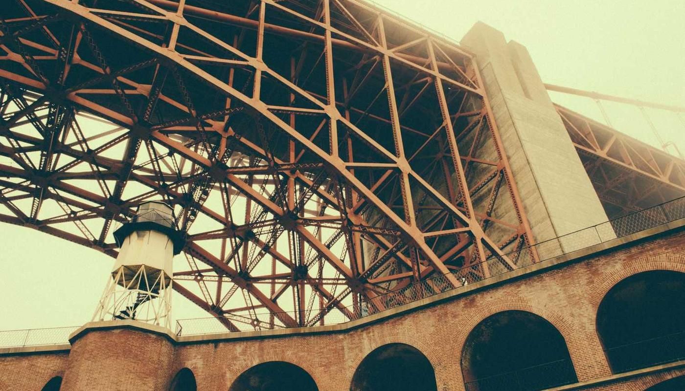 A striking view of a steel truss bridge with a brick archway and a small lighthouse-like tower beneath it, captured in warm, vintage tones.