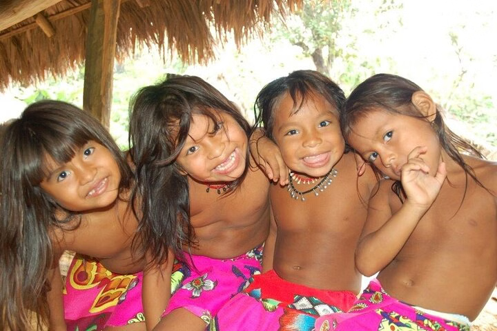 Four Embera children smile and pose together under a thatched roof during a cultural village day trip from Panama City, wearing colorful clothing and surrounded by nature.