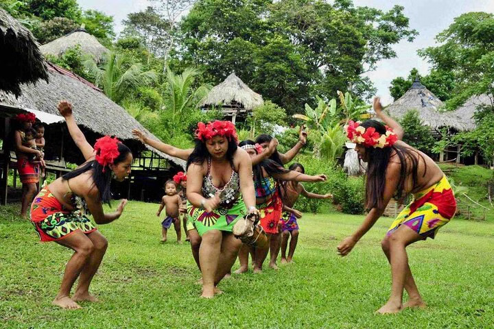 Embera women performing a traditional dance in a tropical village, wearing colorful attire and floral headpieces, surrounded by lush greenery and thatched-roof huts.