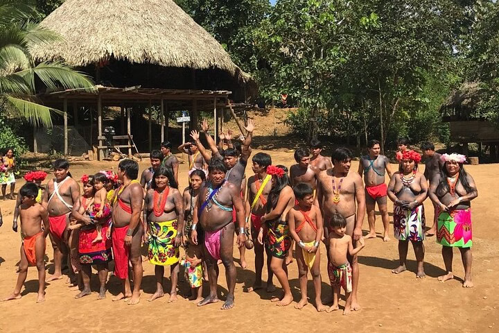 Group of Embera people in traditional attire posing in their village near Panama City, surrounded by lush rainforest and a thatched-roof hut.