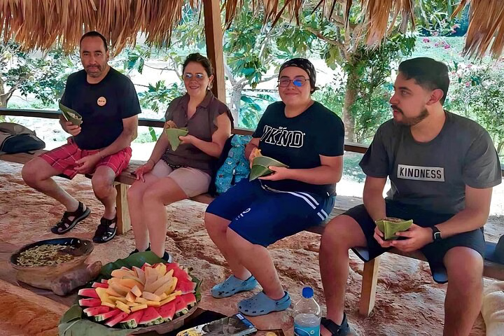 Four travelers enjoy a traditional Embera meal at a thatched-roof hut during a day trip from Panama City, surrounded by lush greenery.