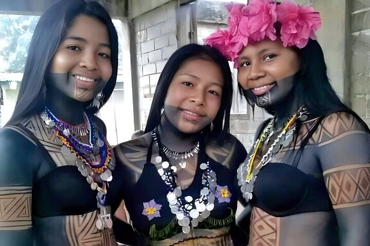 Three Embera women wearing traditional body paint, metal necklaces, and flower headpieces, smiling during a cultural tour in Panama City.