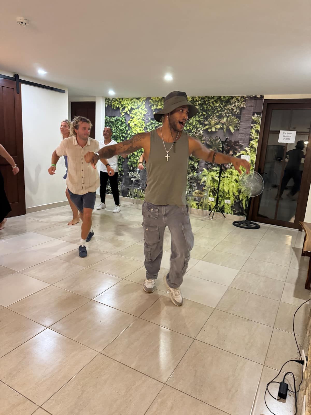 A man leads a salsa dance class in a modern room with a green living wall, surrounded by enthusiastic participants.