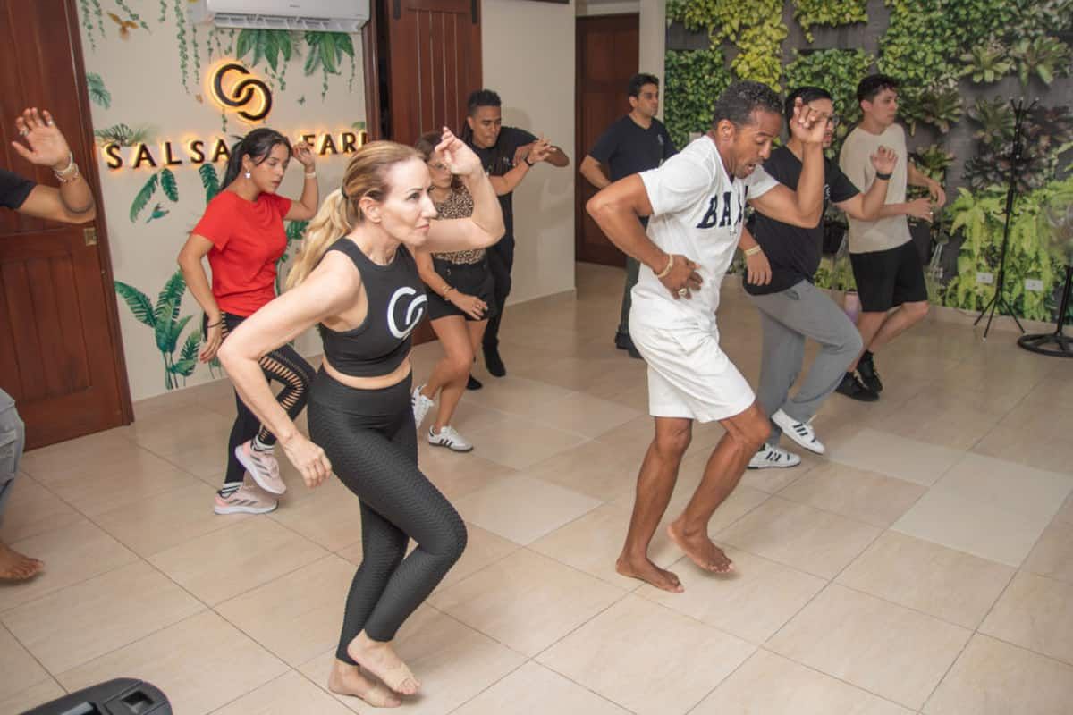 Group learning salsa dance in a modern studio with greenery wall and 'Salsa Safari' branding, led by an instructor in black workout attire.