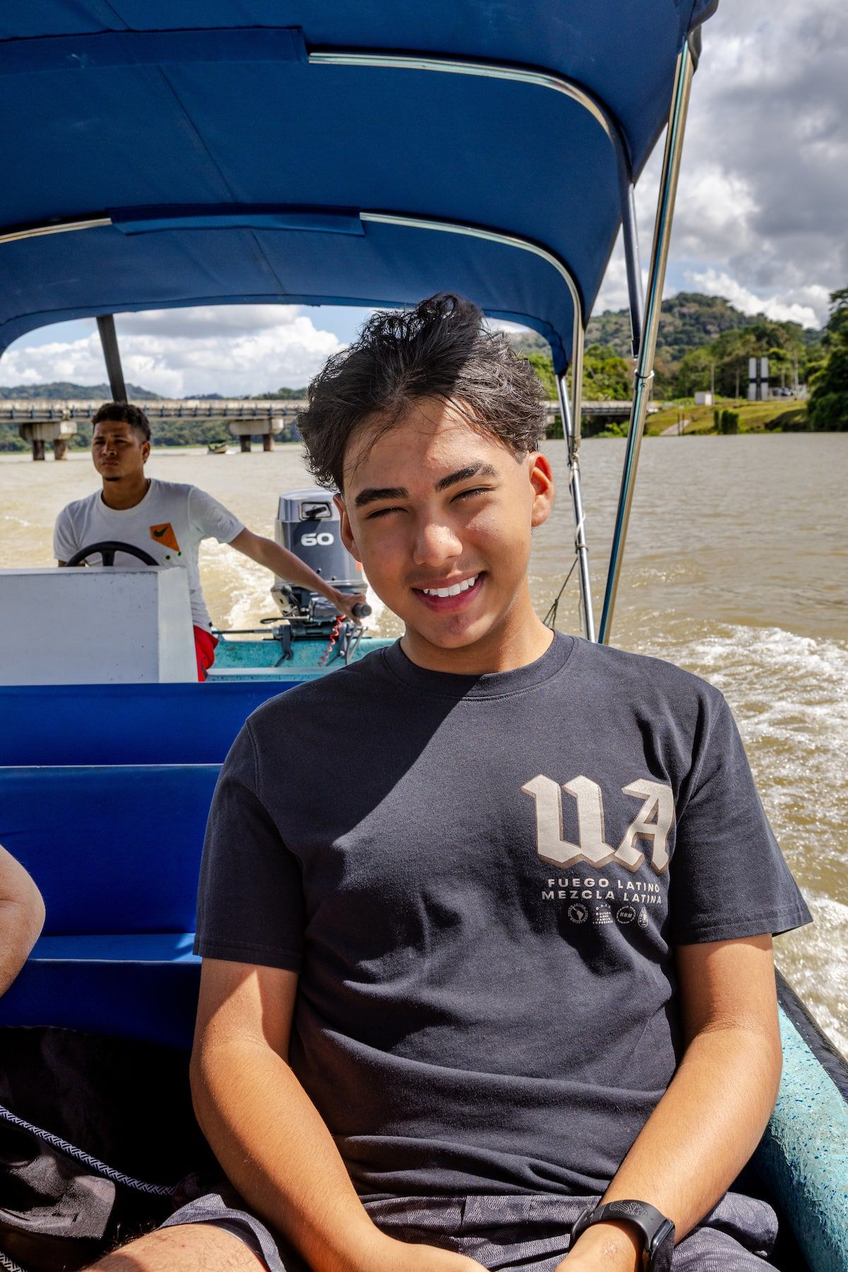 Jeune homme souriant lors d'une visite en bateau sur le lac Gatun à Panama City, avec des collines verdoyantes et un pont en arrière-plan.