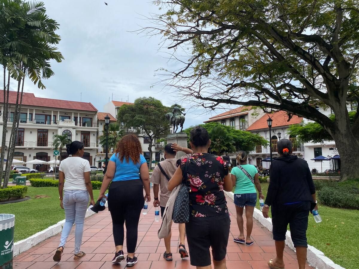 Tourists walking through the historic plaza of Casco Viejo, Panama City, with colonial buildings and a central statue in the background.