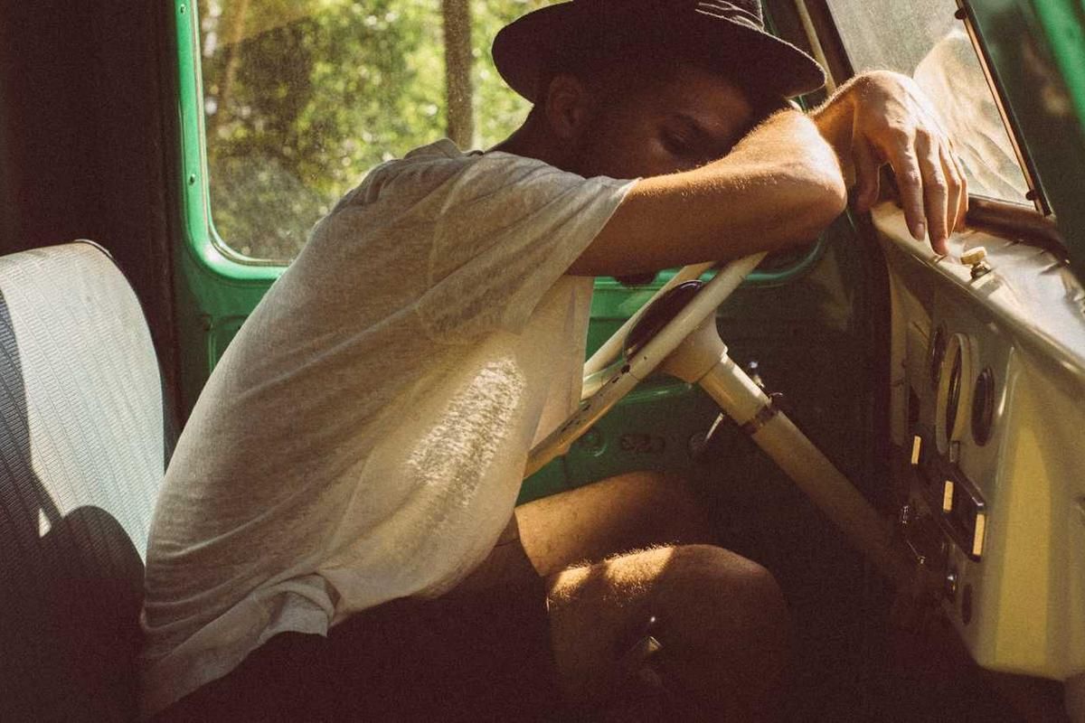 A man rests his head on the steering wheel of a vintage vehicle during a sunny afternoon, evoking a sense of nostalgia and adventure.