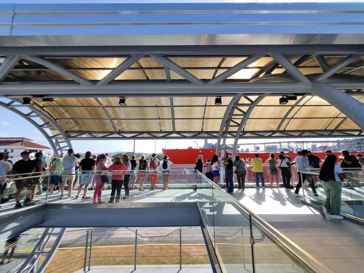 Tourists observe the Panama Canal from a modern viewing platform under a metal and glass canopy, with a red banner in the background.