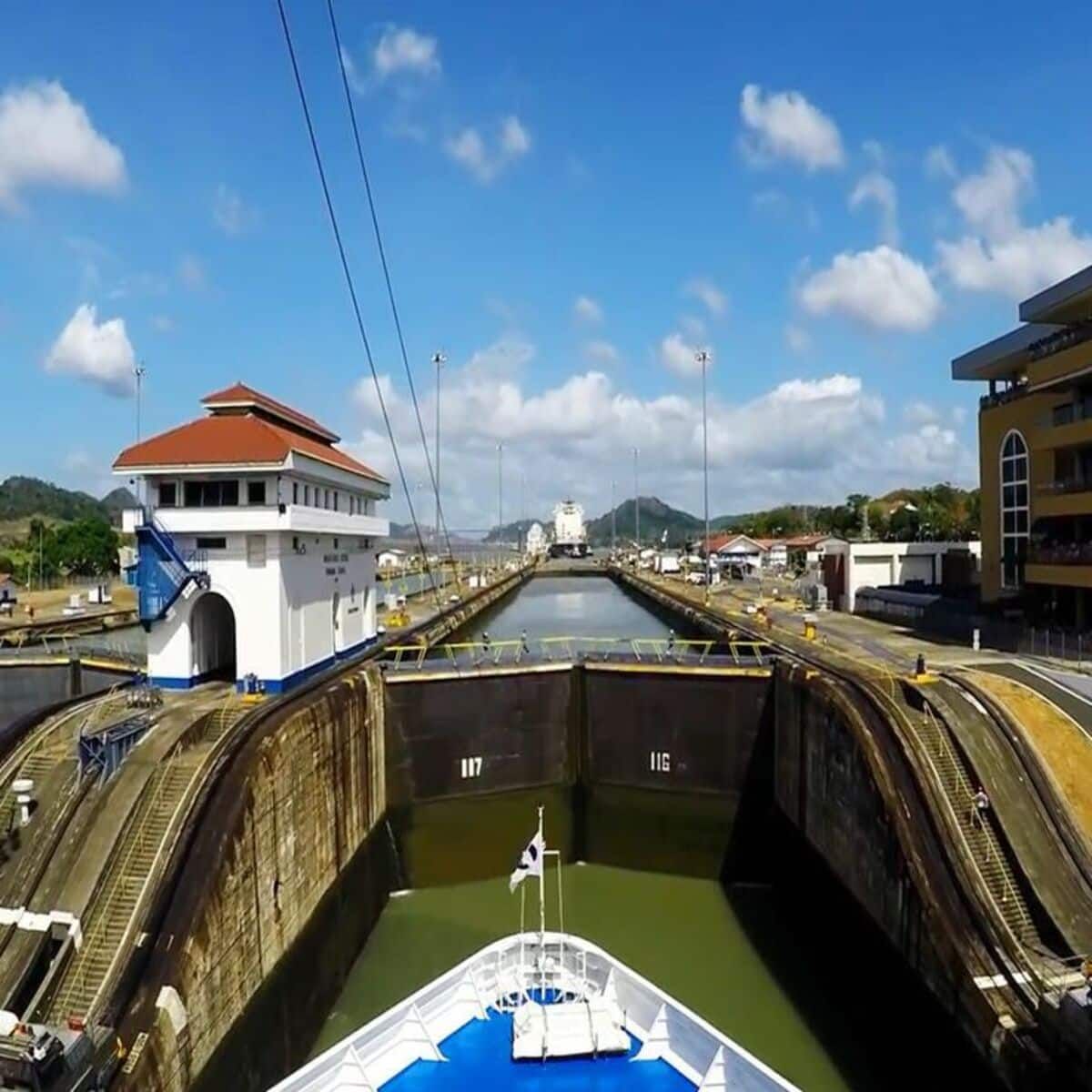 Um barco entra em uma câmara de eclusa no Canal do Panamá, com uma torre de controle e infraestrutura ao redor visíveis sob um céu ensolarado.
