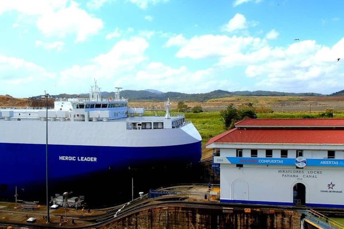 A large cargo ship named 'Heroic Leader' passes through the Miraflores Locks of the Panama Canal, with the iconic control building in the foreground under a sunny sky.