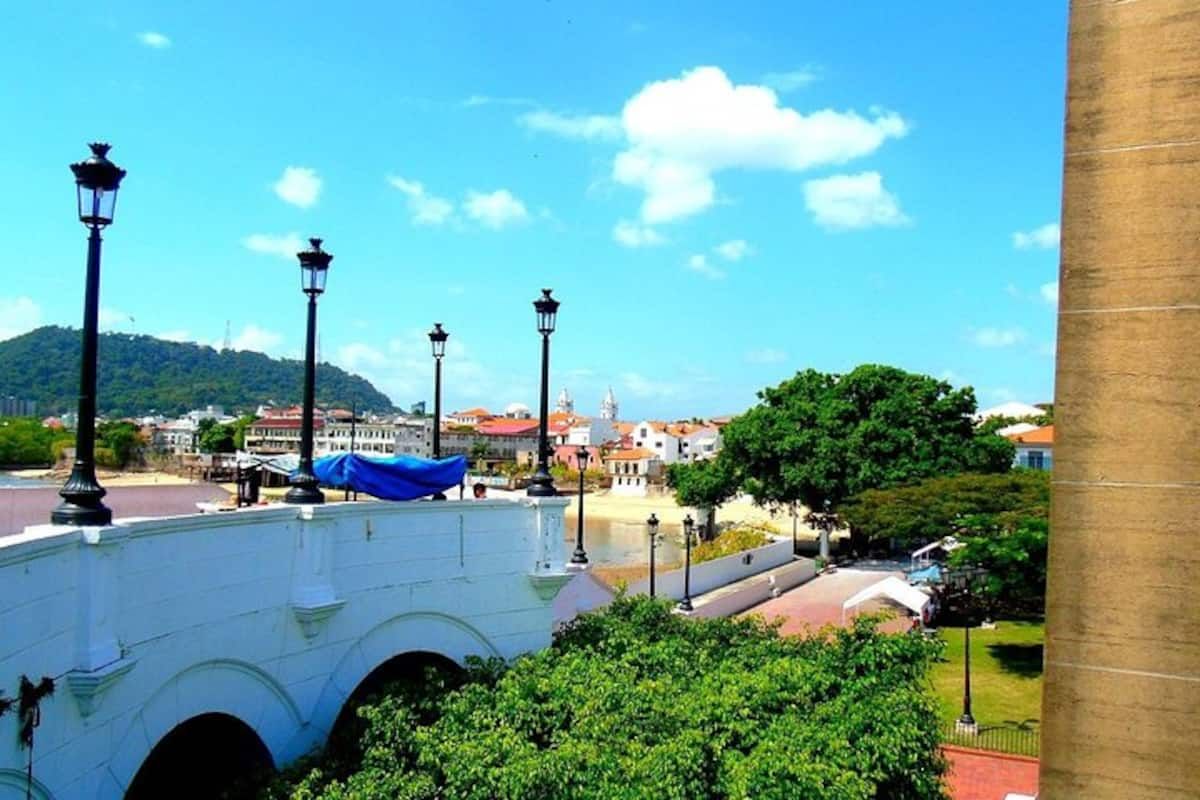White arched bridge with black lampposts overlooks the Miraflores Locks waterway in Panama City, surrounded by greenery and colonial-style buildings under a clear blue sky.