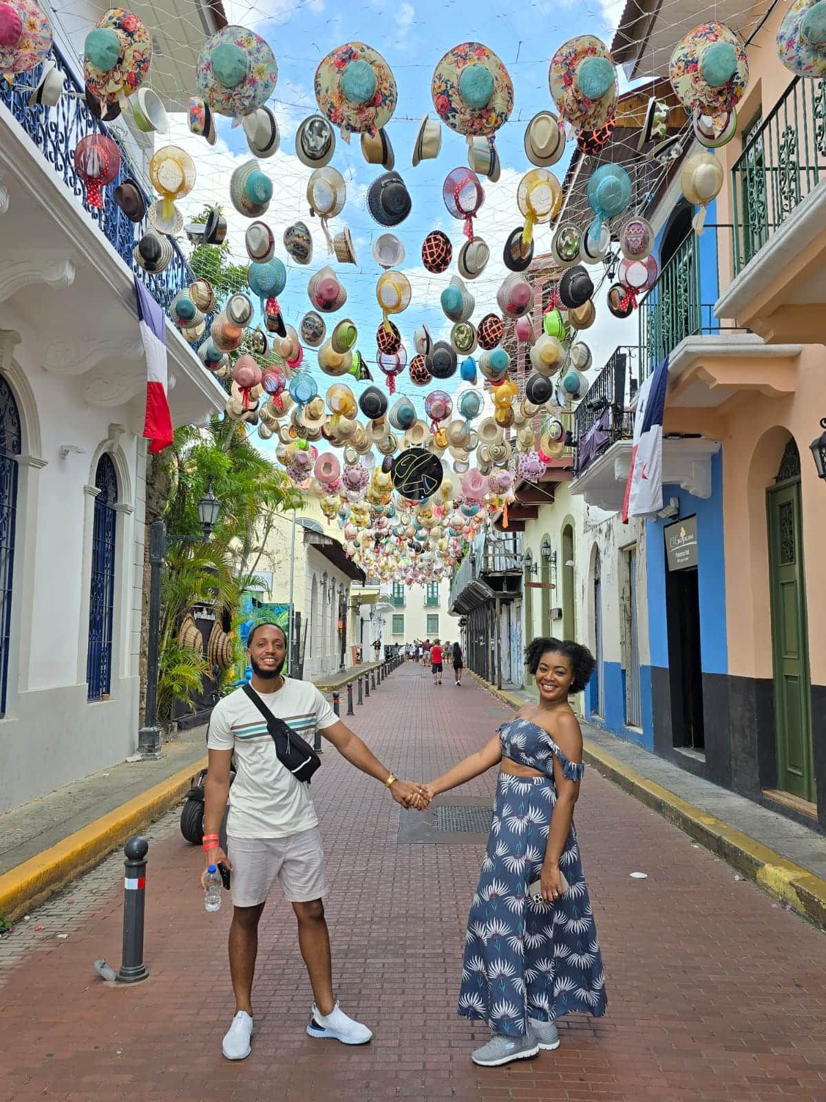 A couple holds hands on a colorful street in Panama City, surrounded by colonial architecture and a canopy of hanging hats during the Panama Canal Visitor Center City Tour.