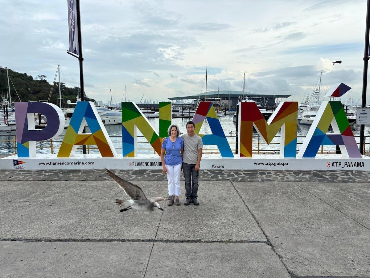 Couple posant devant le coloré panneau 'PANAMA' à Flamenco Marina, avec une mouette volant à proximité et des bateaux en arrière-plan.