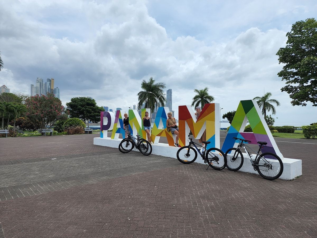 Three people posing with bicycles in front of a colorful 'PANAMA' sign during a bike tour in Panama City, showcasing the city's modern skyline and tropical greenery.