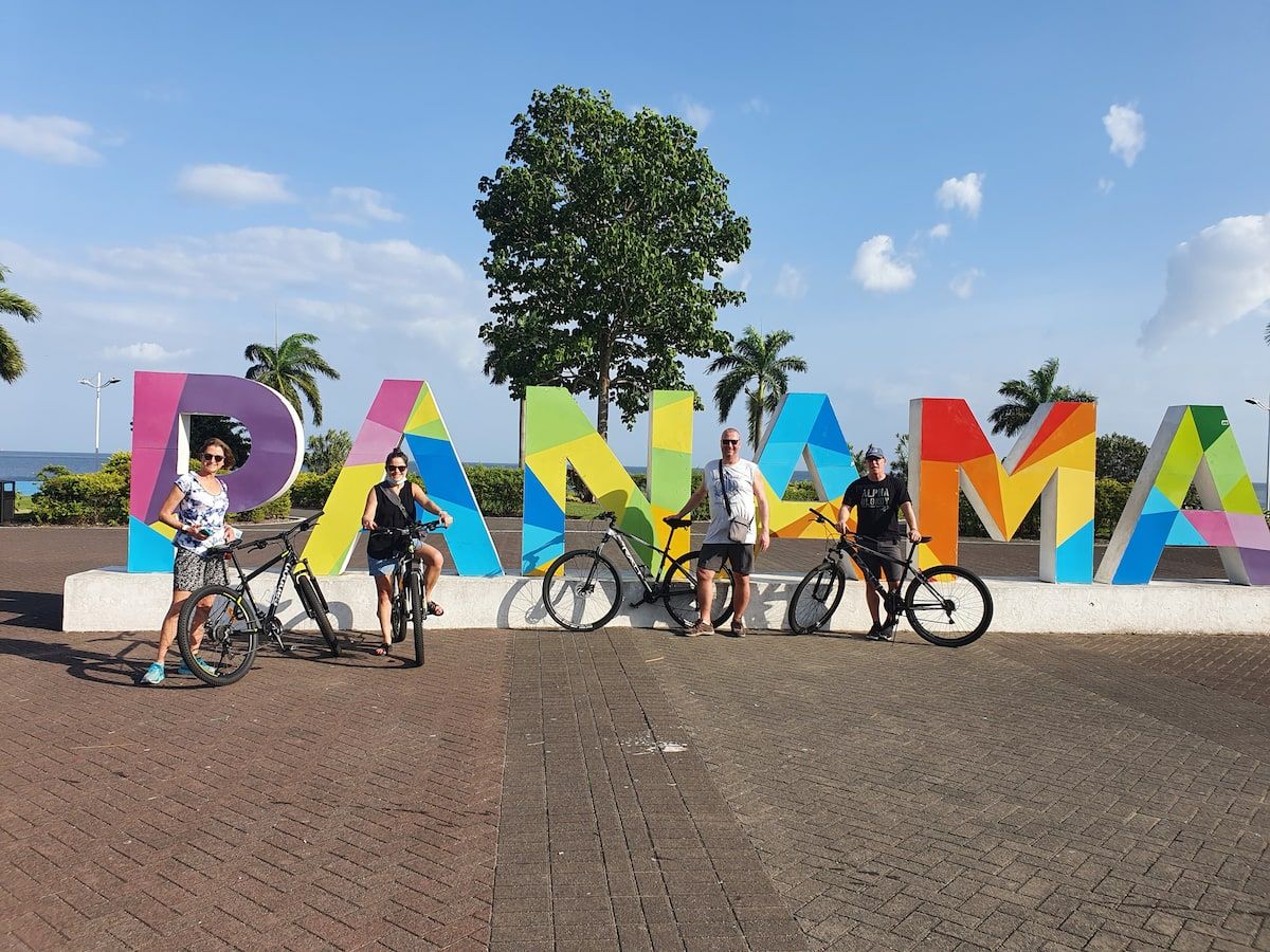 Four cyclists pose in front of a colorful 'PANAMA' sign during a scenic bike tour in Panama City, capturing vibrant skyline views and historic landmarks.