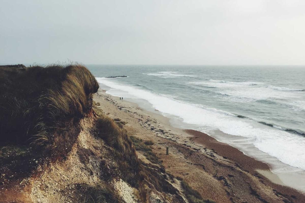 Cliffside beach with dune grass overlooking ocean waves; two people walking along the shore under an overcast sky.