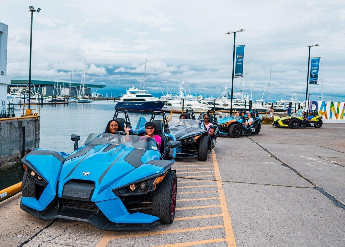 Tourists enjoying a sightseeing tour in blue Polaris Slingshots along the Amador Causeway in Panama City, with a marina and boats in the background.