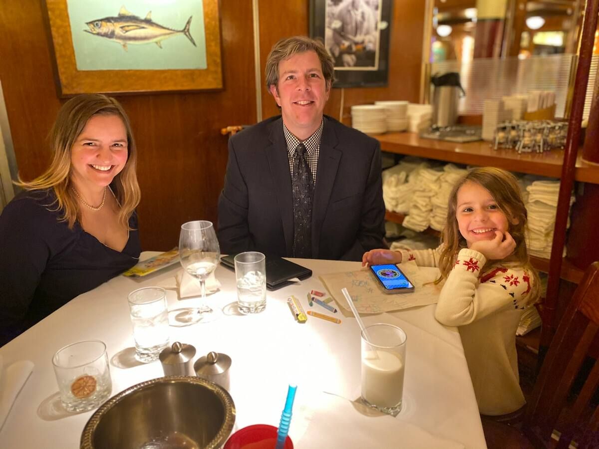 A family of three smiles at the camera while enjoying a meal at a cozy restaurant in Panama City, with drinks and a smartphone on the table.