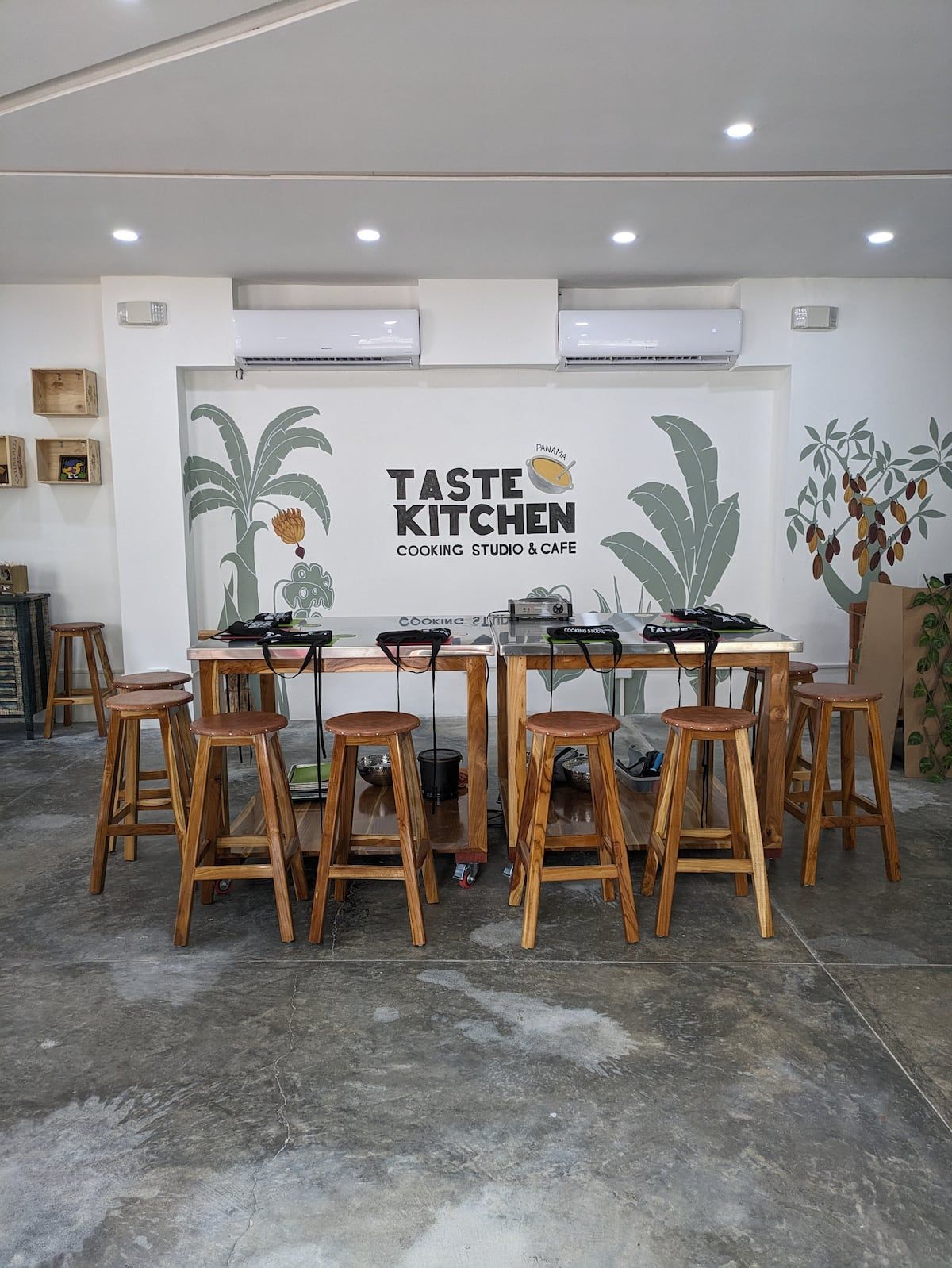 Interior of Taste Kitchen Cooking Studio & Café in Casco Viejo, Panama City, featuring wooden tables and stools set up for a cooking class with tropical plant murals on the walls.