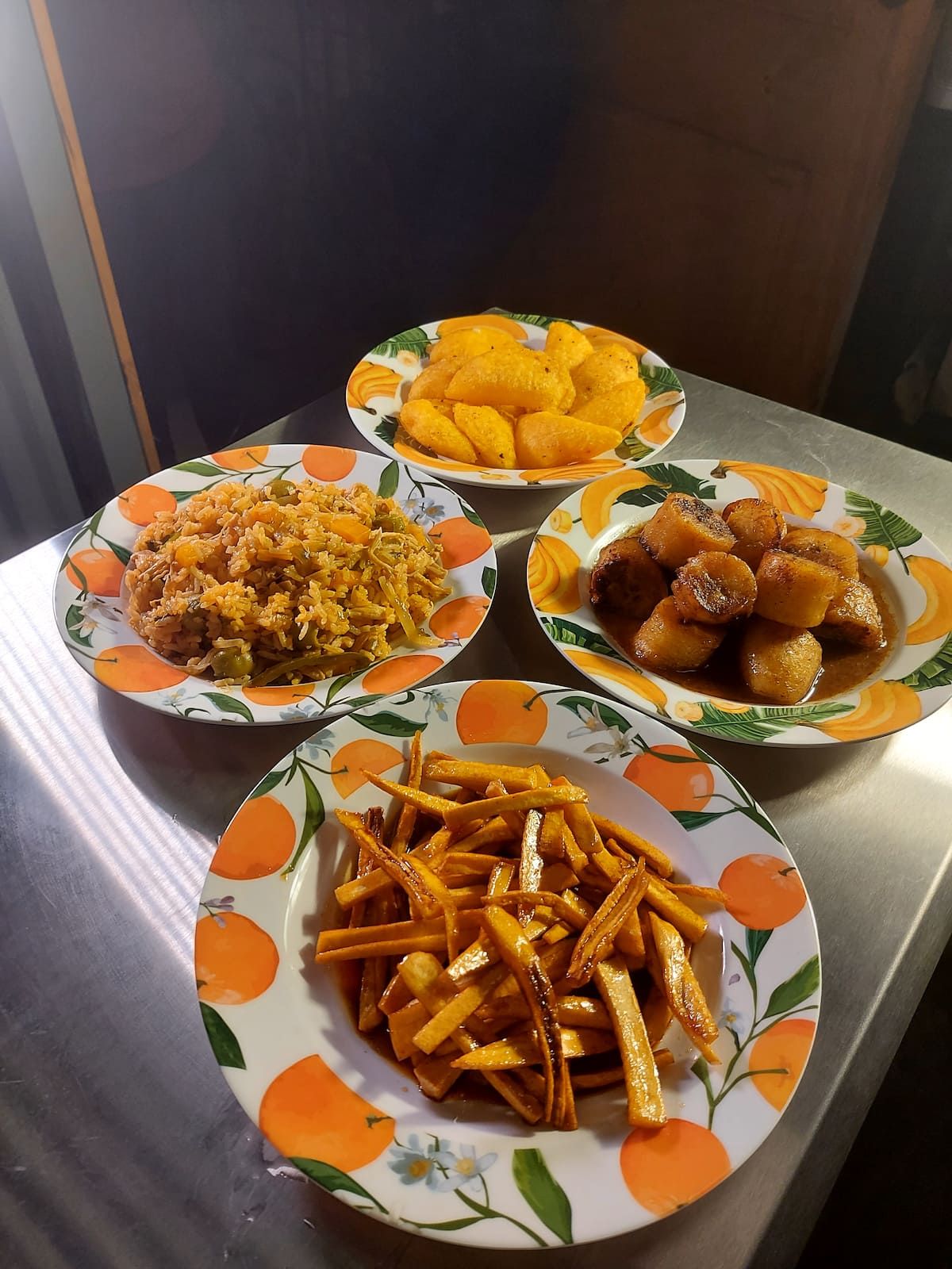 Four traditional Panamanian dishes served on orange-patterned plates, including fried plantains, rice with vegetables, fried yuca, and sweet plantain slices, presented on a metal table with natural lighting.