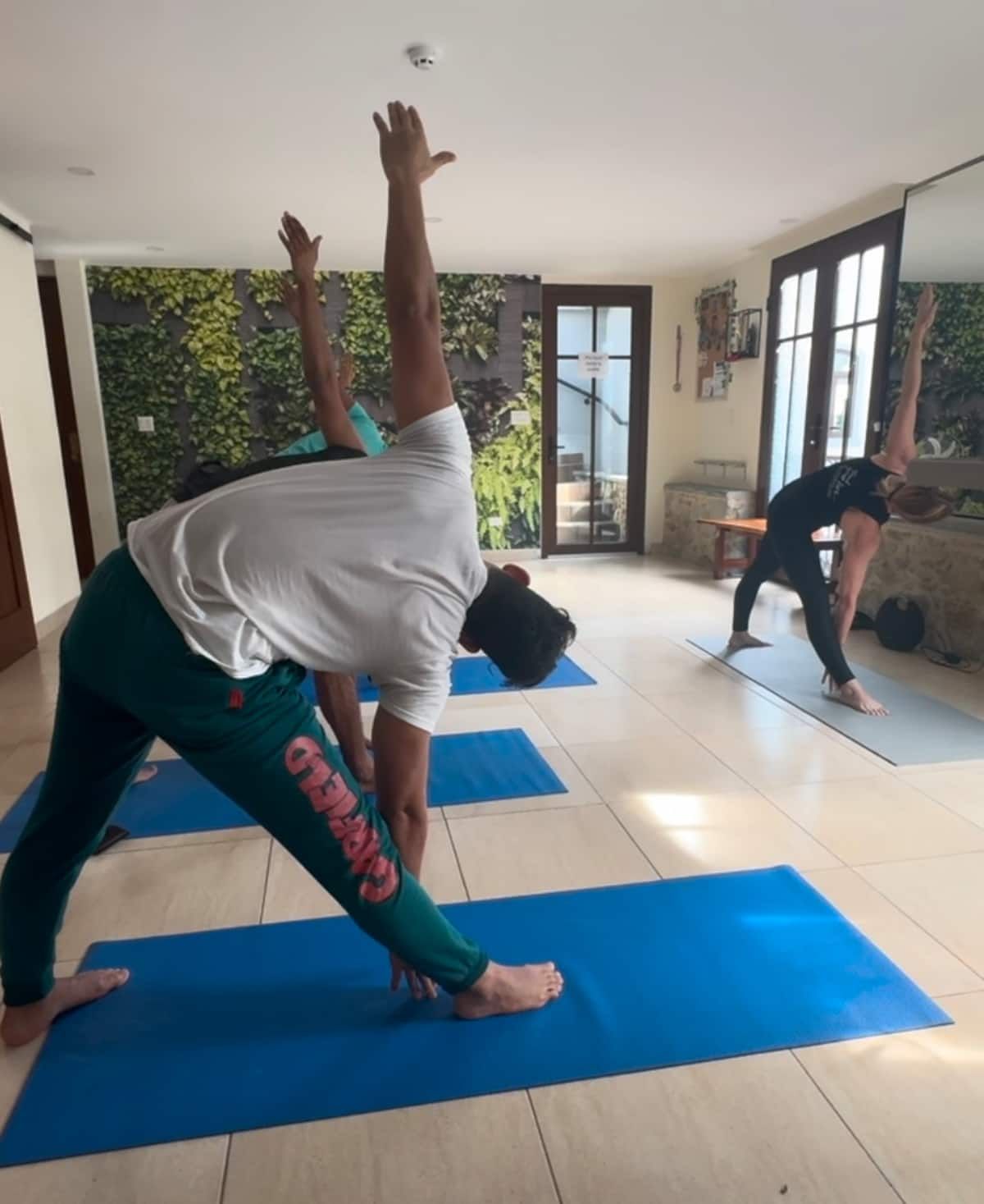 Participants practicing yoga in a bright studio with a green living wall, wearing white shirts and green pants on blue mats.