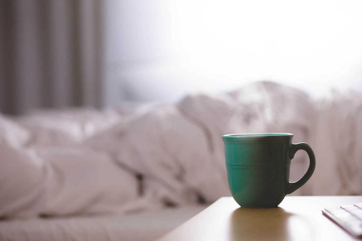 A teal coffee mug sits on a wooden table in a softly lit room with white bedding, evoking a sense of calm before an adventurous day.