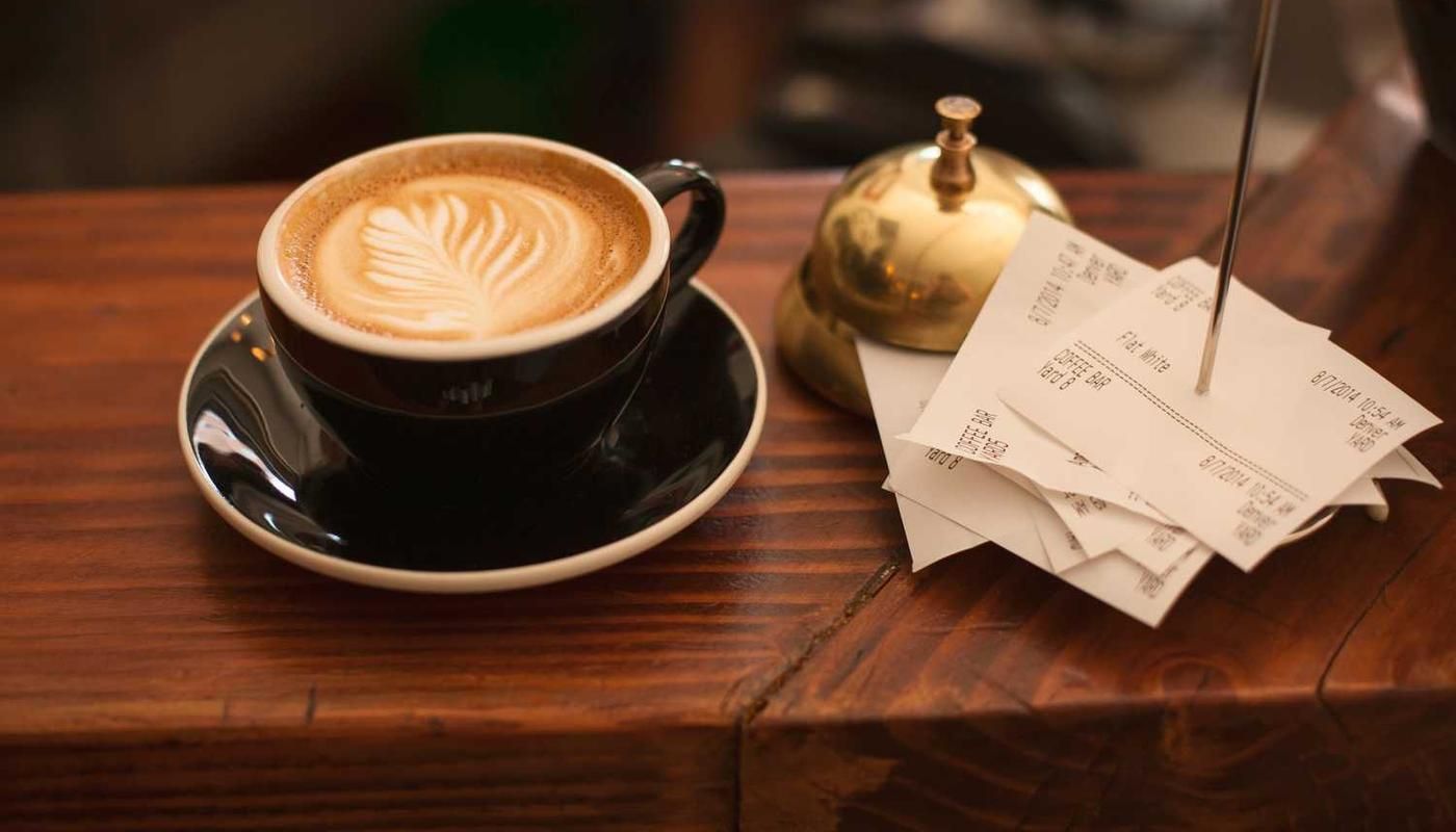 A latte with leaf-shaped milk foam art sits on a wooden counter next to receipts and a golden service bell in a cozy café setting.